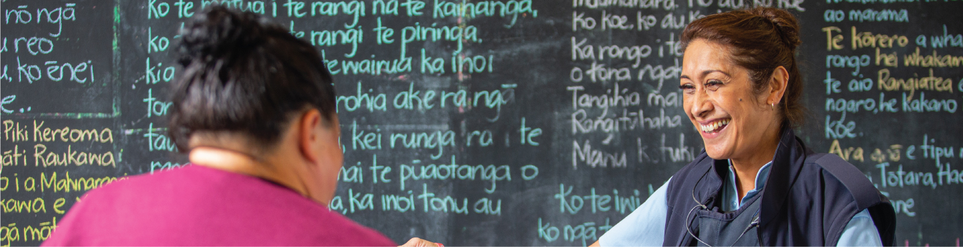 Corrections Officer smiles and talks with a woman, with a blackboard with te reo written in chalk in the background