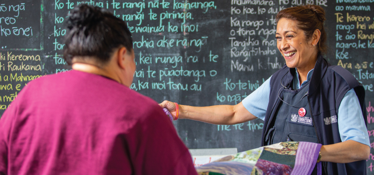 Corrections Officer smiles and talks with a woman, with a blackboard with te reo written in chalk in the background
