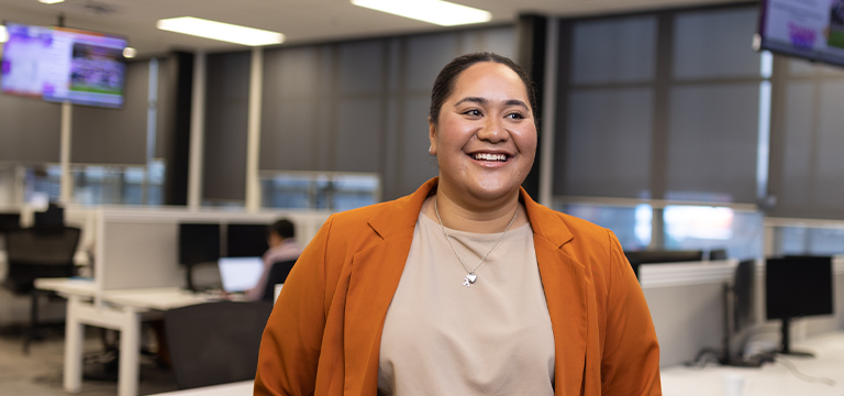 Operations Adviser stands smiling in office, with screens hanging from the ceiling and rows of desks in the background