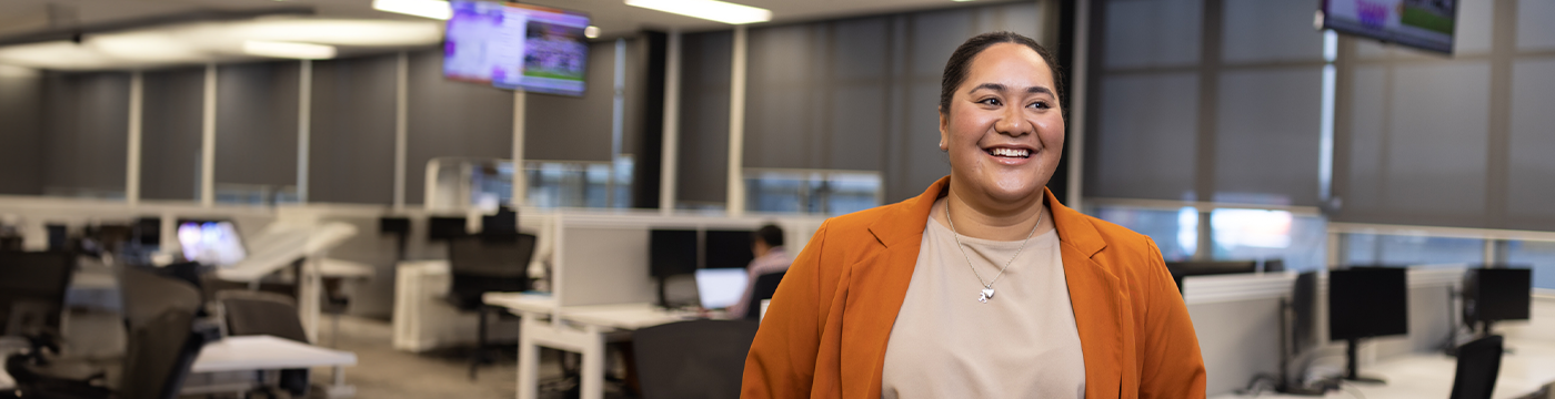 Operations Adviser stands smiling in office, with screens hanging from the ceiling and rows of desks in the background