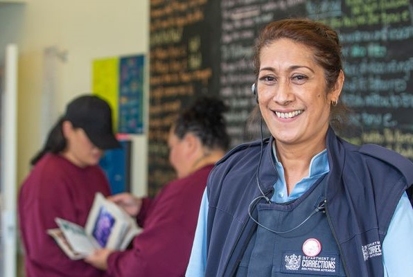 Corrections Officer stands smiling with blackboard with te reo written on in the background