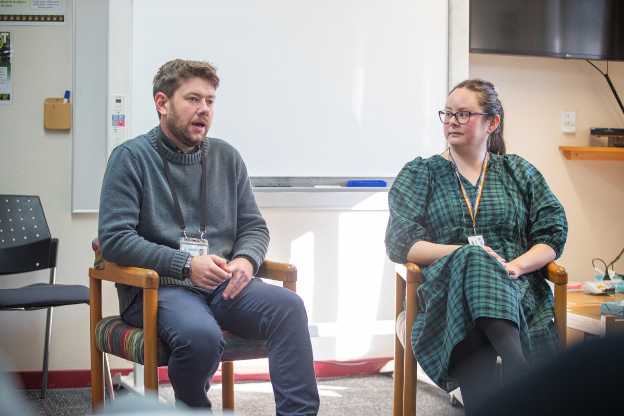 Psychologists sit in chairs as part of a group. One psychologist is talking while the other listens intently