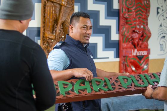 Corrections Officer holds a sign with te reo on and engages with two men, with Māori artwork in the bavkground